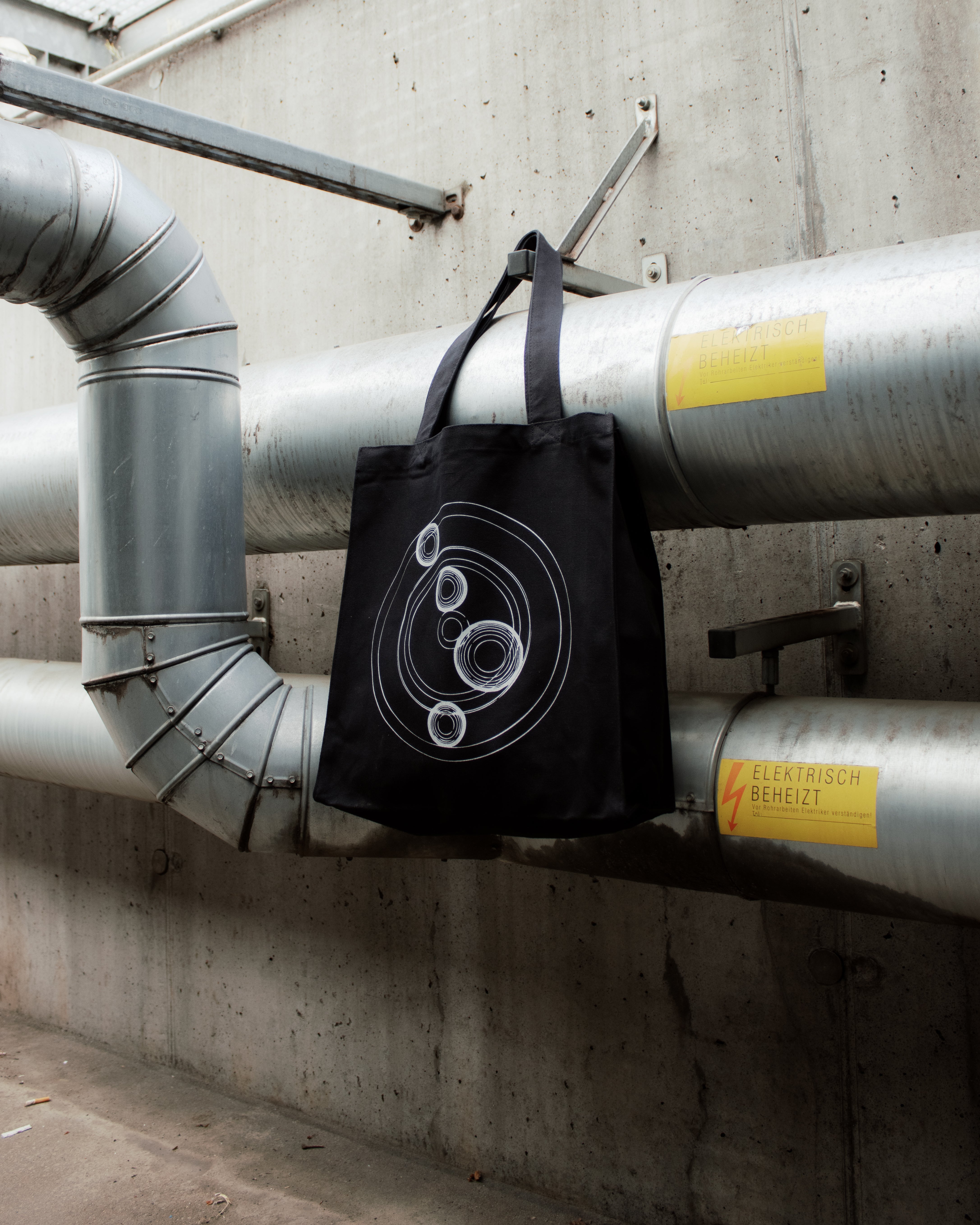 Black tote bag with geometric design hanging on industrial pipes against a concrete wall. "Cocoon Icon"