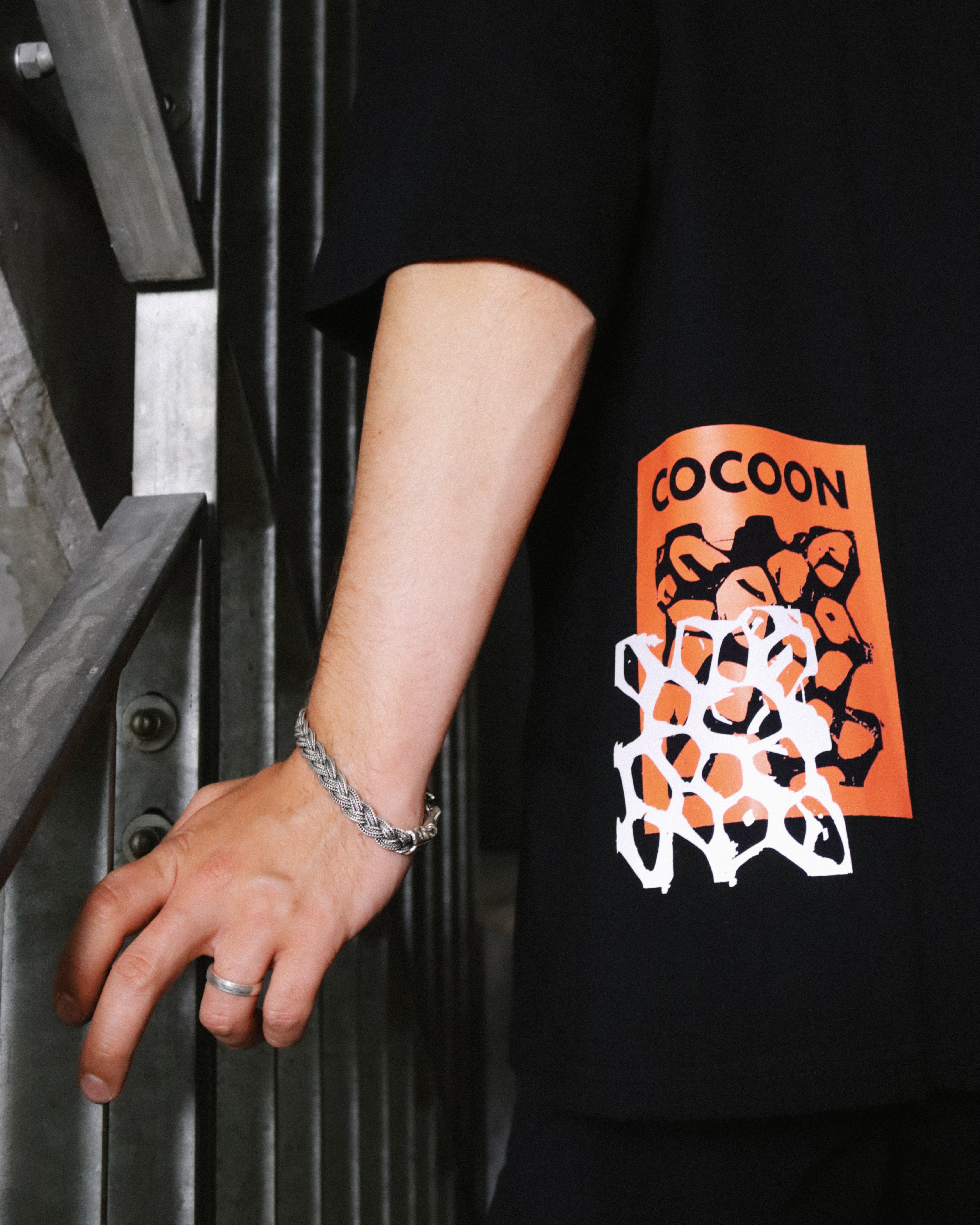 A close-up of a person wearing a black oversized t-shirt with a graphic print. The graphic features an orange square containing black abstract shapes and the word 'COCOON', overlaid with a white geometric pattern. reflective honeycomb graphic detail