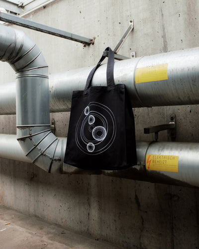 Black tote bag with geometric design hanging on industrial pipes against a concrete wall. "Cocoon Icon" 