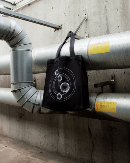 Black tote bag with geometric design hanging on industrial pipes against a concrete wall. "Cocoon Icon" 