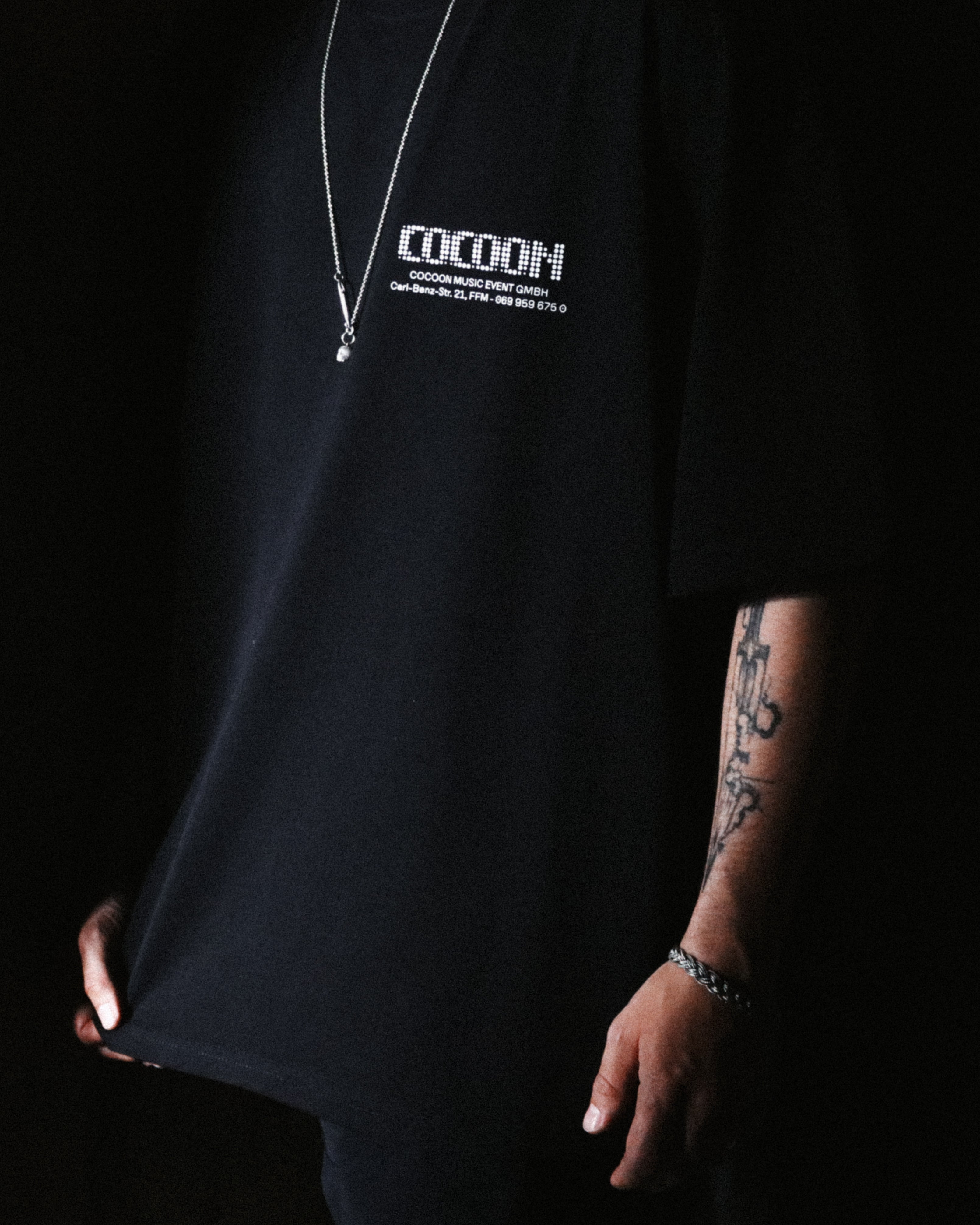 A person wearing an oversized black t-shirt with white 'COCOON' text printed on the chest against a dark background. Address T-shirt