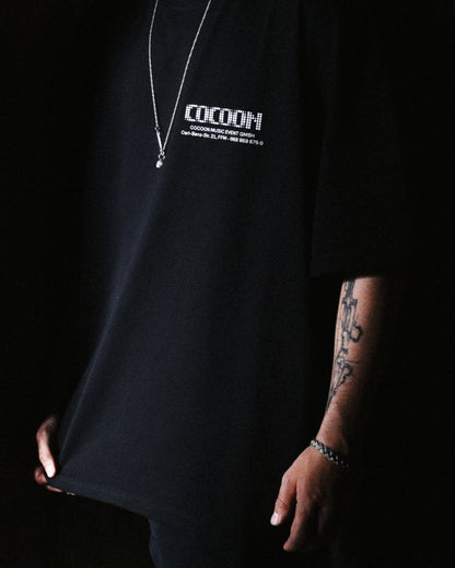 A person wearing an oversized black t-shirt with white 'COCOON' text printed on the chest against a dark background. Address T-shirt